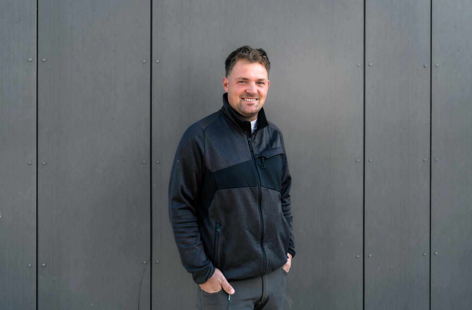 Ein Mann in dunkler Kleidung steht lässig vor einer grauen Metallwand, seine Hände ruhen in der Hosentasche, das Gesicht ist zensiert.