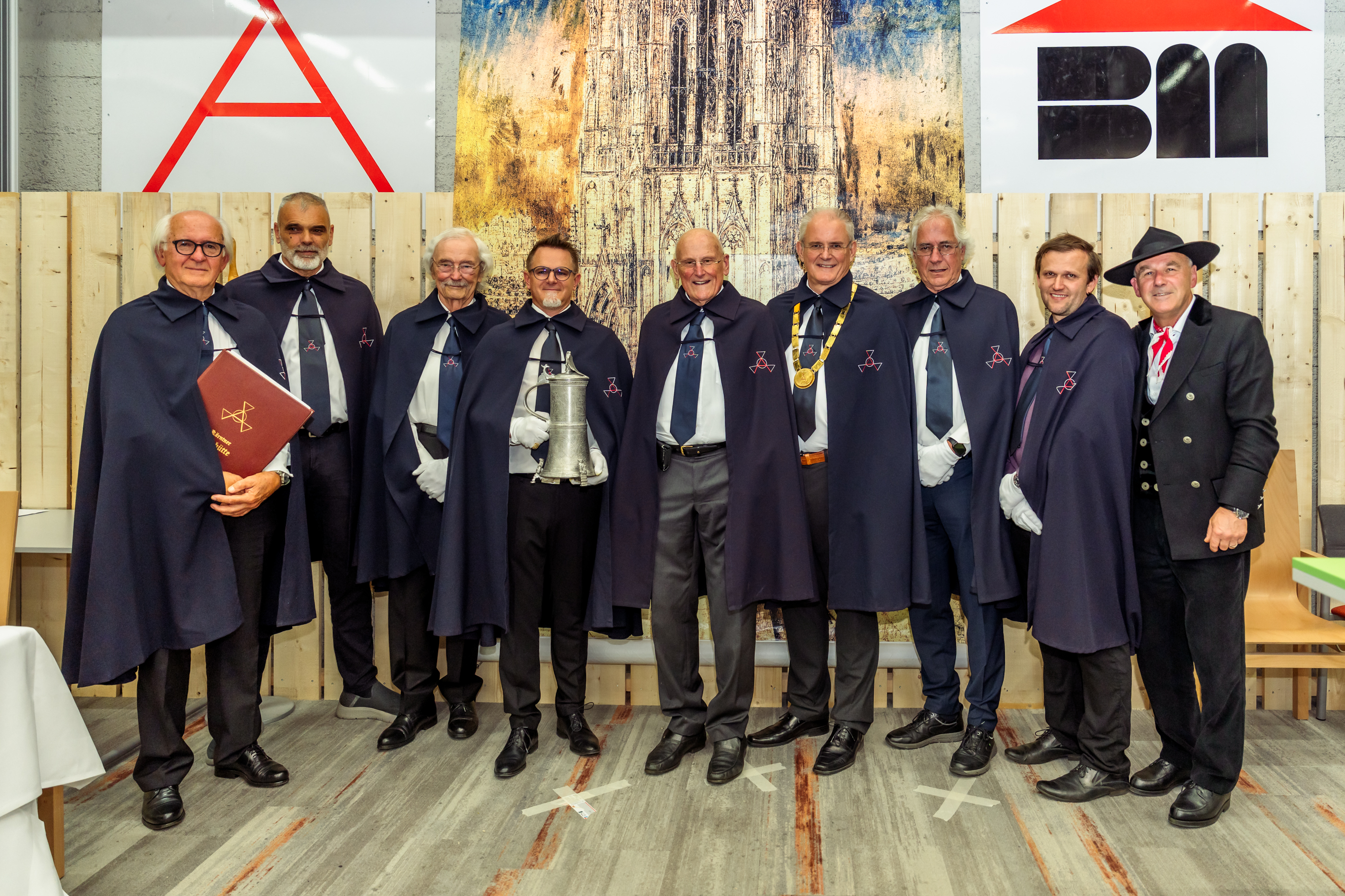 Gruppenfoto von zehn Personen in festlicher Zeremonienkleidung vor einer dekorierten Holzwand. Einige tragen Insignien oder halten symbolische Gegenstände, darunter eine Urkunde und einen Zinnkrug.