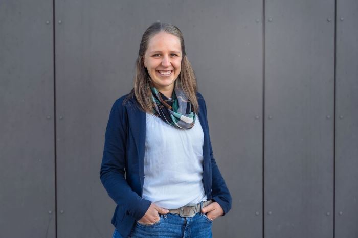 Eine Frau in Freizeitkleidung steht vor einer grauen Wand. Sie trägt eine blaue Jacke, ein weißes Shirt, eine Jeans und einen gemusterten Schal. Ihre Hände sind entspannt in den Hosentaschen.