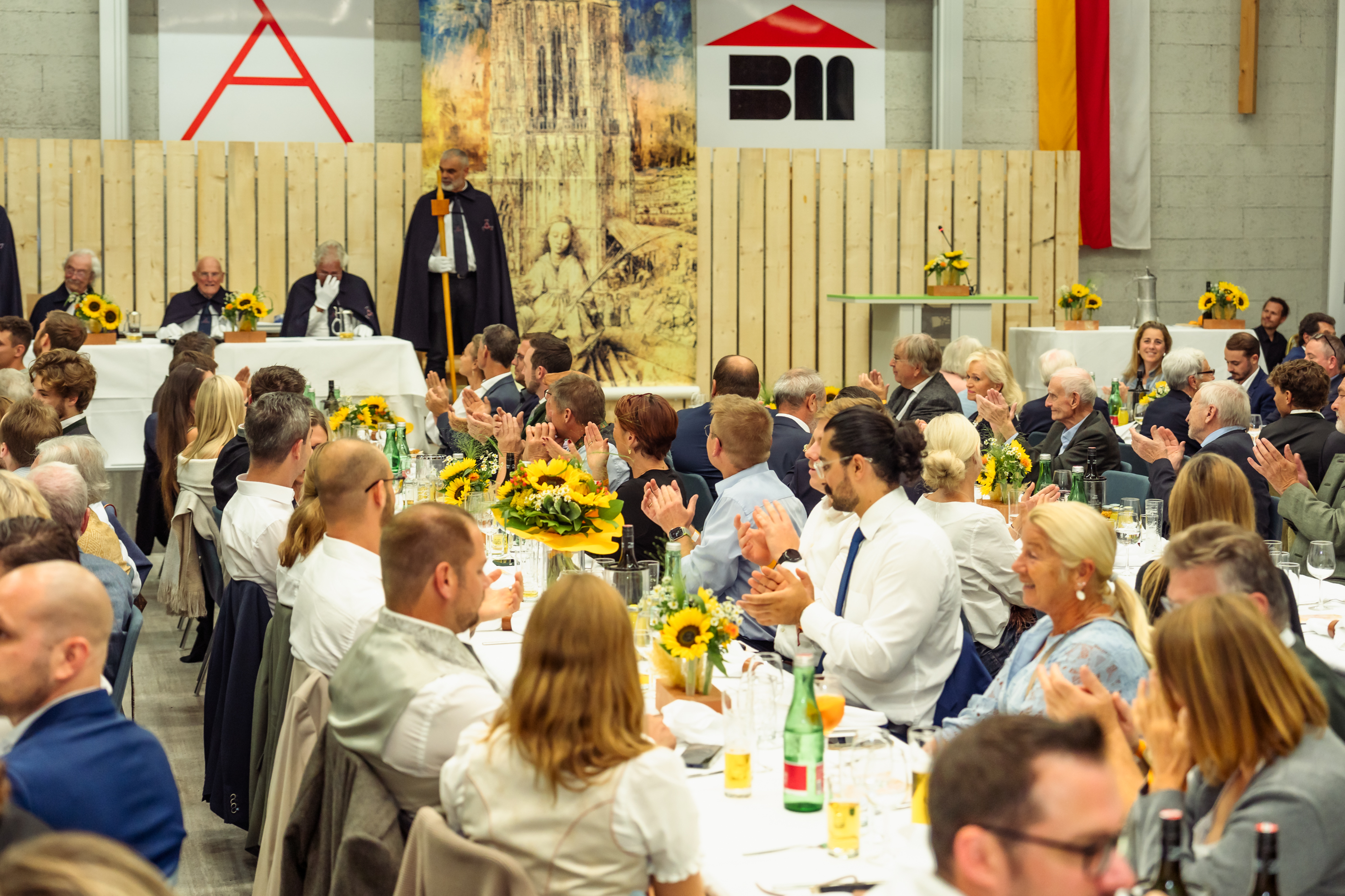 Festveranstaltung in einem großen Saal: Zahlreiche Gäste sitzen an festlich gedeckten Tischen mit Sonnenblumenarrangements und applaudieren, während eine Person in dunklem Umhang vor dem Ehrentisch steht.
