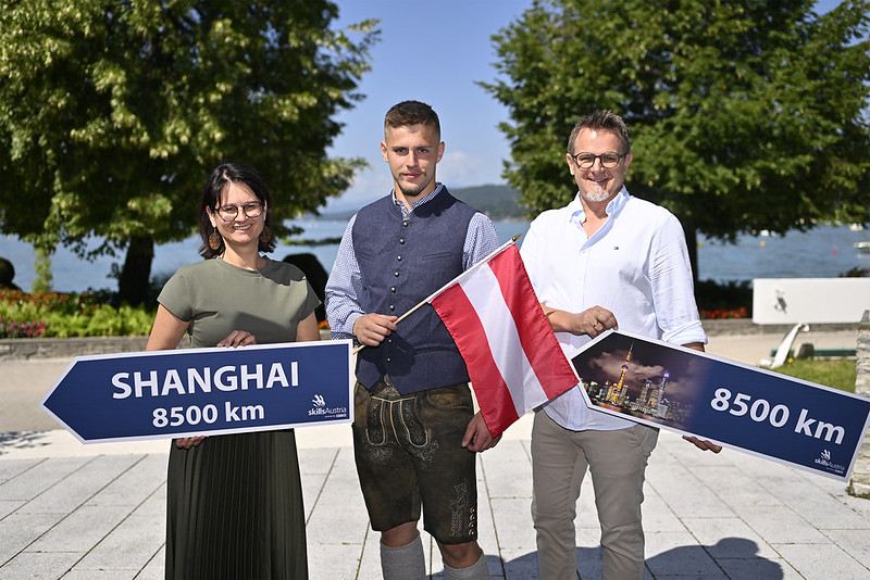 Österreichischer Teilnehmer mit Begleitpersonen präsentiert vor einer Seepromenade die österreichische Flagge und Wegweiser mit der Aufschrift „Shanghai 8500 km“.