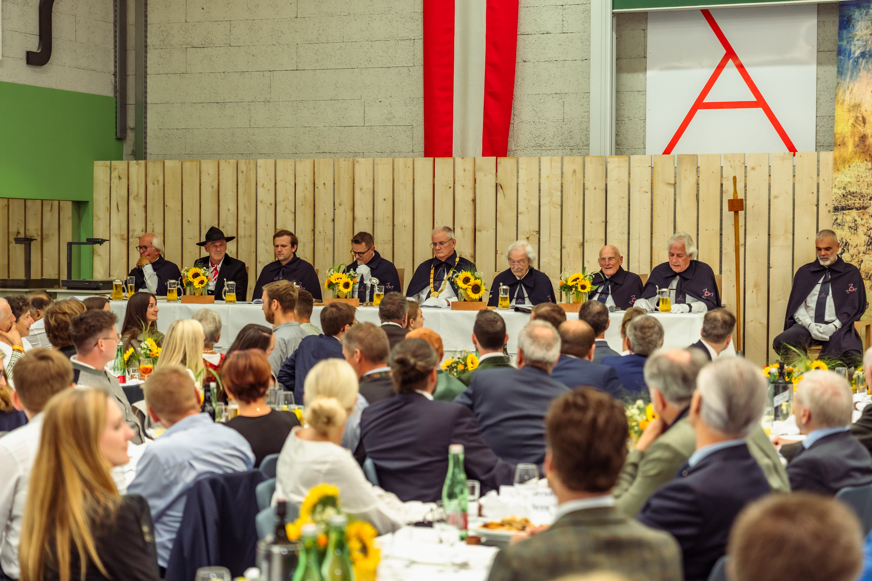 Festliche Veranstaltung mit zahlreichen Gästen an gedeckten Tischen. Am Ehrentisch sitzen mehrere Personen in dunkler Zeremonienkleidung vor einer Holzkulisse mit österreichischer Flagge und Sonnenblumenarrangements, während das Publikum der Veransta