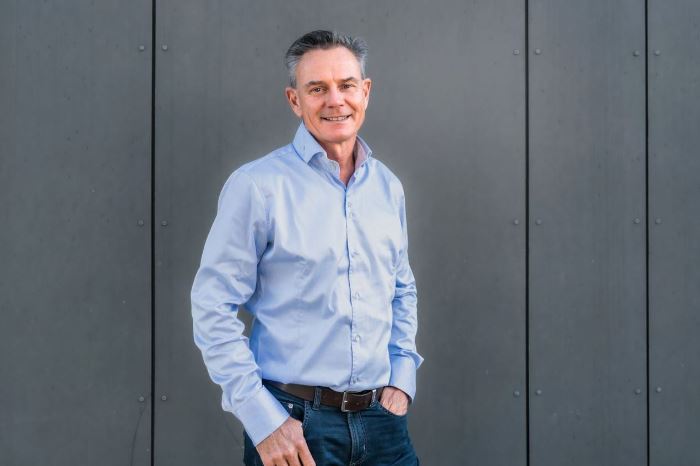 Ein Mann trägt ein hellblaues Hemd und Jeans, steht entspannt mit einer Hand in der Tasche vor einer grauen Wand. Sein Gesicht ist unkenntlich gemacht.