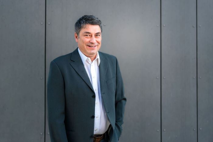 Ein Mann in eleganter, dunkler Kleidung steht vor einer grauen Wand. Die Kleidung wirkt formell und eignet sich für einen beruflichen Kontext.